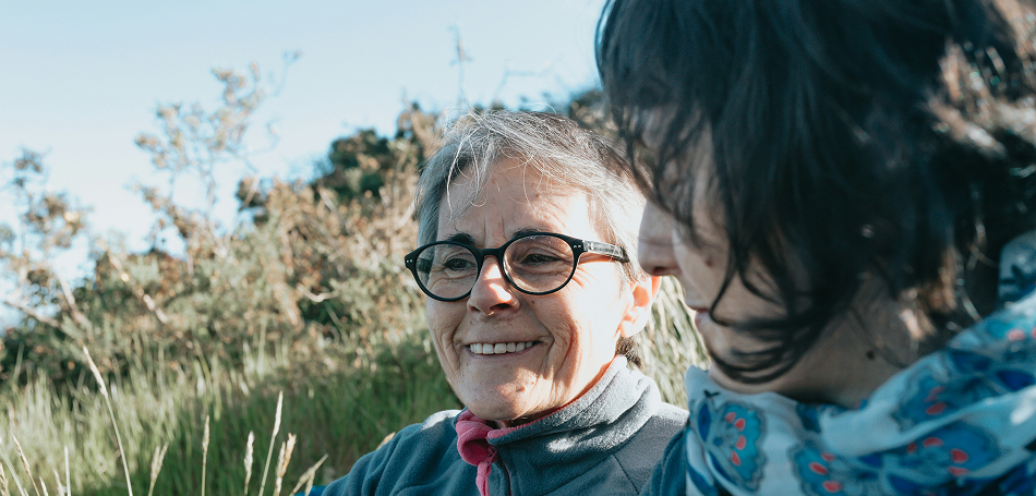 Two women smiling together outdoors in a grassy natural setting.