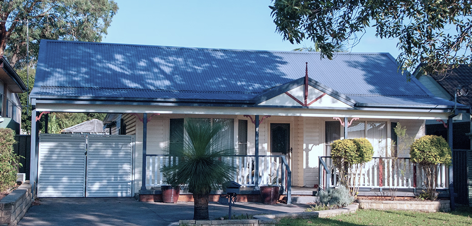 The front of a single-storey Australian weatherboard home with a verandah, corrugated iron roof, and a grass tree in the garden.