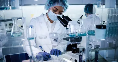 Laboratory scientist reviewing testing data while working with a microscope in a diagnostic laboratory, surrounded by laboratory glassware and analytical equipment used to support quality control, calibration verification and accurate diagnostic testing.