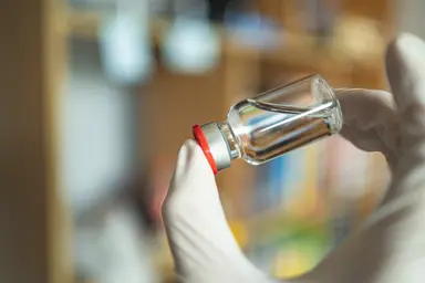 Gloved laboratory hands holding a glass vial, representing virus-like particle development, vaccine research and diagnostic reagent production.