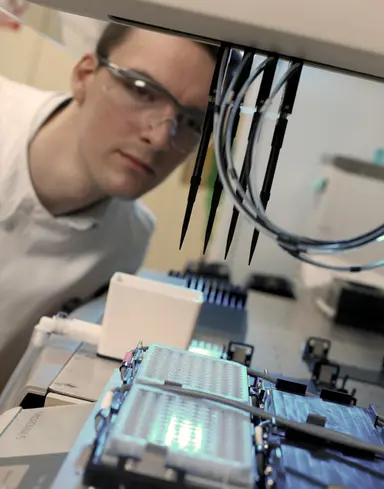Close-up of an automated laboratory pipetting system processing microplates while a laboratory professional monitors the workflow, representing precision liquid handling, assay development, laboratory automation and high-throughput testing.