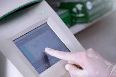 Close-up of a PCR thermal cycler touchscreen displaying an amplification protocol, with a gloved hand adjusting temperature and cycling parameters during assay setup.