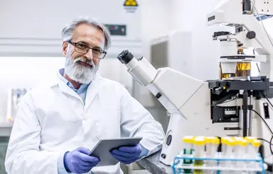 Laboratory professional reviewing testing data on a tablet beside a microscope and sample tubes, representing quality control practices, third-party QC materials and performance monitoring in a clinical laboratory.