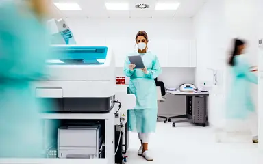 Laboratory professional using a tablet alongside automated analytical instruments in a clinical testing laboratory, representing informatics software, digital quality control data management and secure laboratory workflow monitoring.
