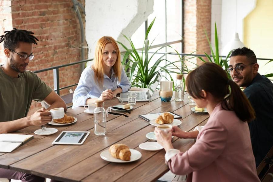 A group of smiling people gather around a table with coffee, pastries, and tablets.