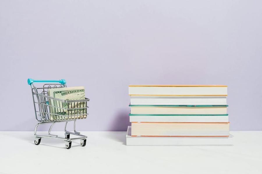 A tiny shopping cart holding dollar bills sits beside a stack of books.