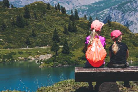 Two children sit on a bench overlooking an alpine lake.
