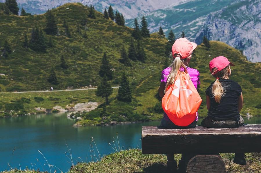 Two children sit on a bench overlooking an alpine lake.