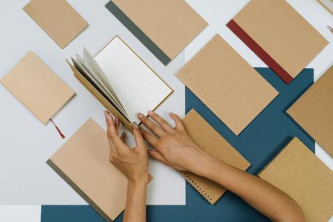 Hands open a brown notebook surrounded by other brown notebooks of various types.