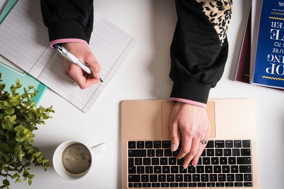 Overhead view of a person's hands—one hand on a laptop keyboard and the other writing in a notebook.