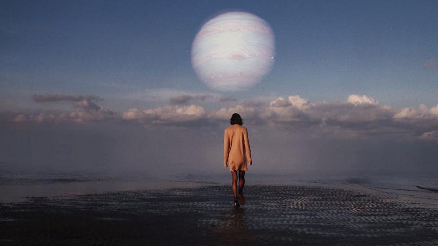 A person stands on a beach with a giant planet looming overhead.