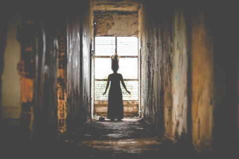 Silhouette of a woman with raised hair standing in front of a window at the end of a rusted, decrepit corridor.