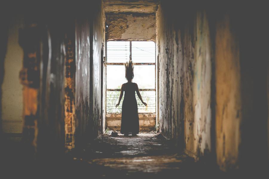 Silhouette of a woman with raised hair standing in front of a window at the end of a rusted, decrepit corridor.