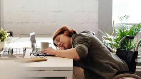 A person in a wheelchair sleeps at a desk beside a computer.