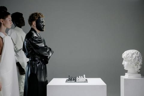 A person with a painted face plays chess against a Grecian sculpture.