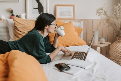 A writer lies on a bed on their stomach, sipping a cup of coffee and looking at a laptop screen.