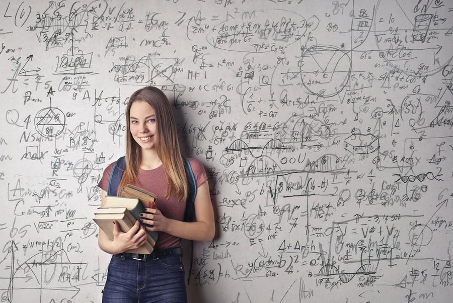 Woman holding books in front of a white wall with scribbles and formulas written on it.