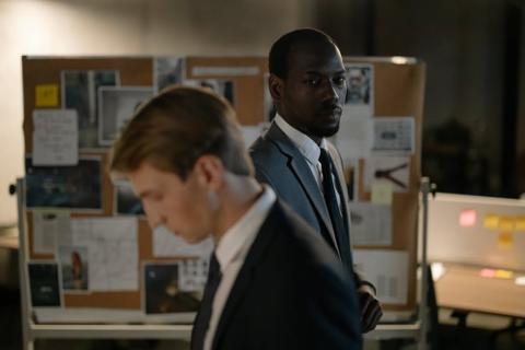 Two detectives stand together facing different directions.