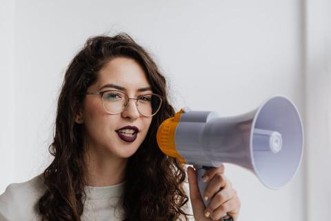 A person with long hair and dark lipstick speaks into a megaphone.
