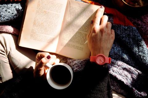 A person's arms with painted fingernails and a pink watch, holding a cup of coffee and an open book.