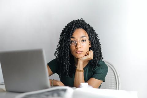 A writer sits beside a laptop, resting their chin in their hand and looking directly at the camera.