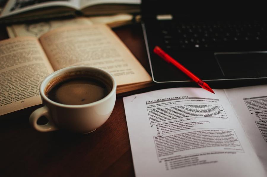A table with a cup of tea, a book, a notebook, a pencil, and a laptop.