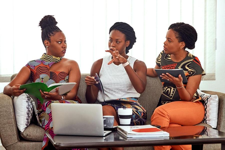 Three people sit on a couch holding notebooks and having an animated discussion. One of them looks annoyed.