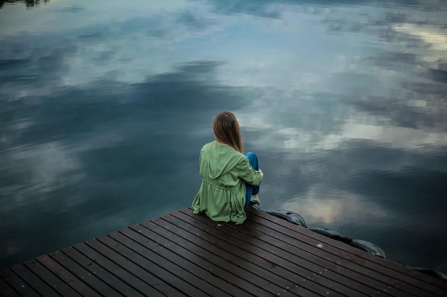 A person in a green jacket and long brown hair sits alone on a dock overlooking a lake.