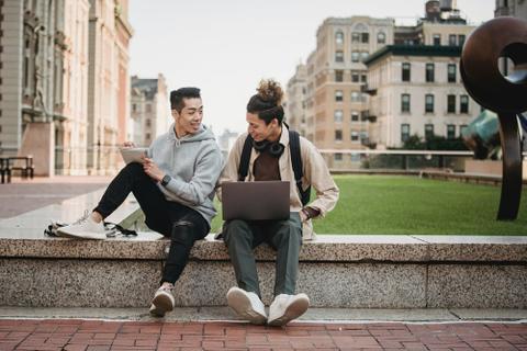 Two young men sit on a short wall, discussing the contents of a notebook.