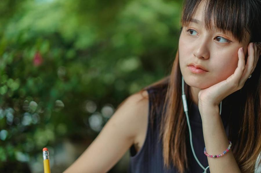 A writer wearing headphones holds a pencil and rests their chin on their hand as they stare off into the distance.