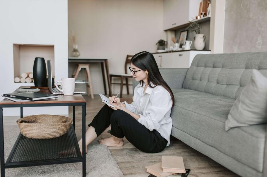 A person sits on the floor between a couch and a coffee table, writing in a notebook.