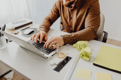 A person types on a laptop next to pieces of crumpled paper.