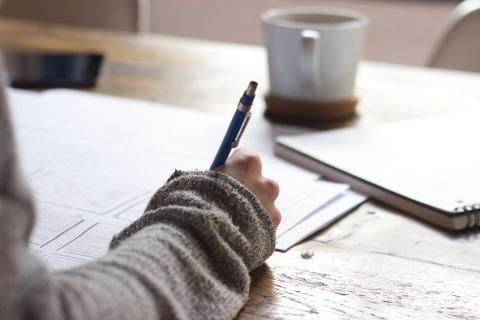 A hand writes in a notebook on a wooden desk.