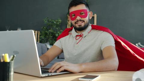 A person in a mask and cape sits at a computer.