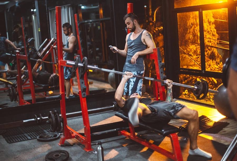 Man struggling to bench press a barbell while someone else stares at their phone.