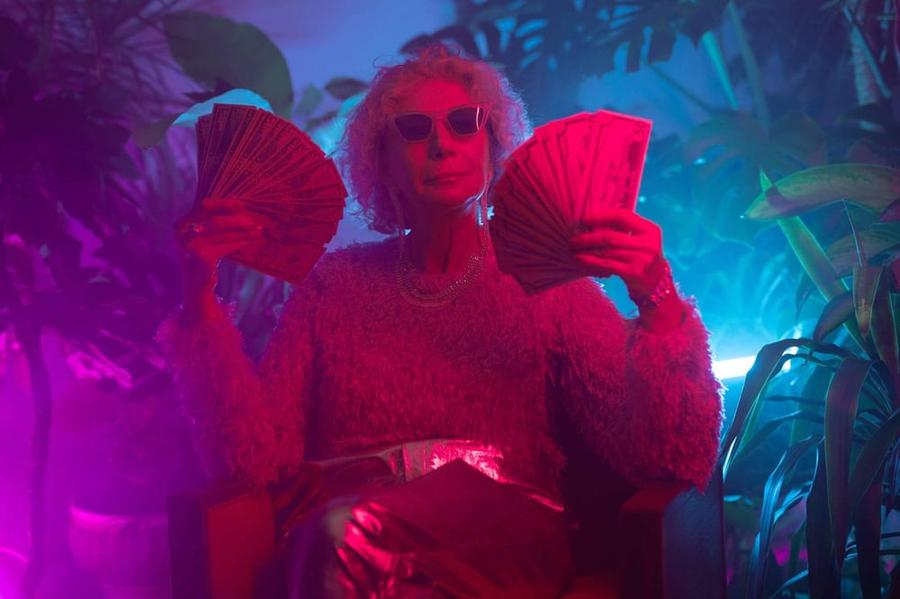 An elderly person wearing sunglasses holds up two fans of cash while basking in multi-colored lights.