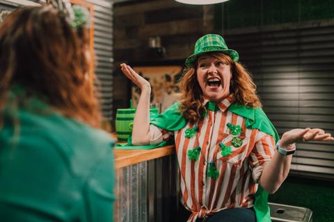 A person in a shamrock necklace and green leprechaun hat smiles and shrugs at another person.