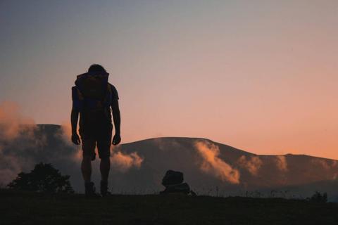Silhouette of a person wearing a backpack and looking out over a mountain range at dusk.