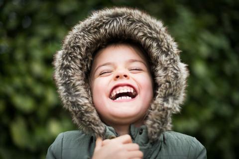 A child in a coat with a fur hood laughs with their head slightly tipped back.