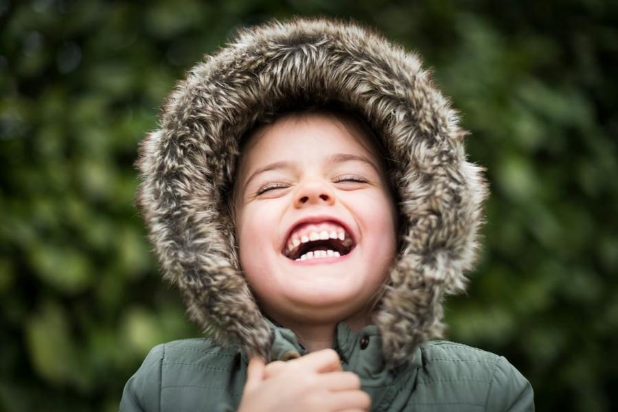 A child in a coat with a fur hood laughs with their head slightly tipped back.