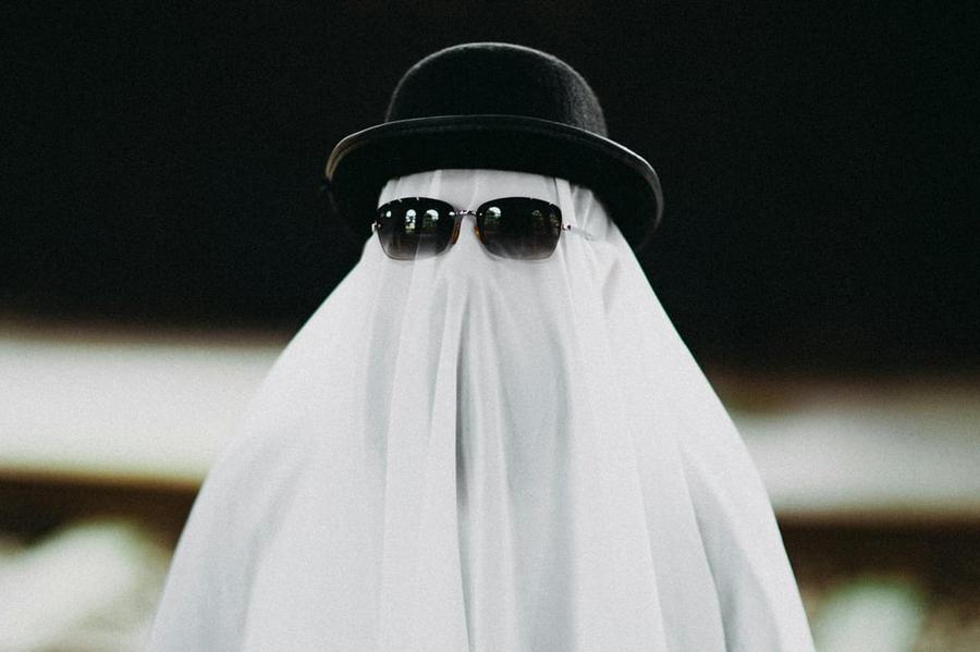 A person with a sheet draped over them like a ghost. They also wear a hat and sunglasses.
