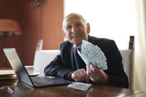 An older man in a suit, sitting in front of an open laptop holding money.