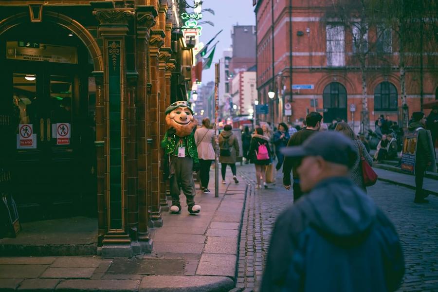 A crowd of people in a busy city street with a person in a leprechaun outfit on the sidewalk.