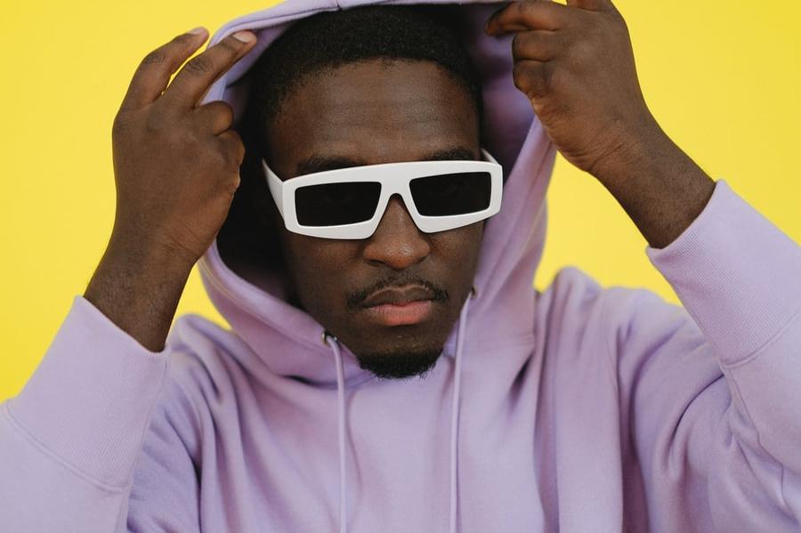 A person wearing dark sunglasses pulls up the hood of their pink hoodie.