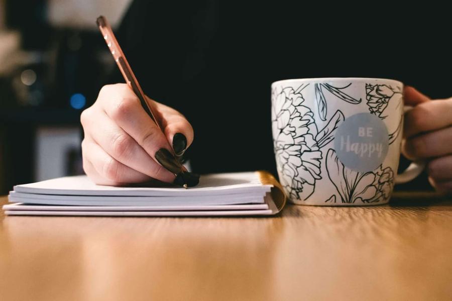 Hand writing in a notebook next to a big coffee mug.