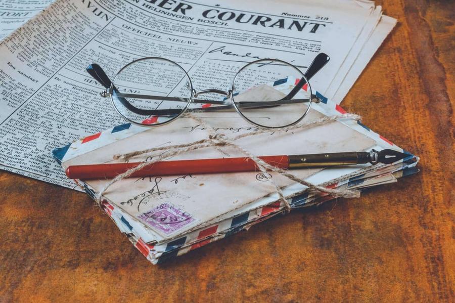 A stack of old envelopes tied together with a fountain pen on top of a newspaper and under a pair of round glasses.