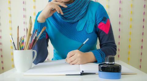 An image from the neck down of a person in a bright blue sweater writing in a blank notebook.