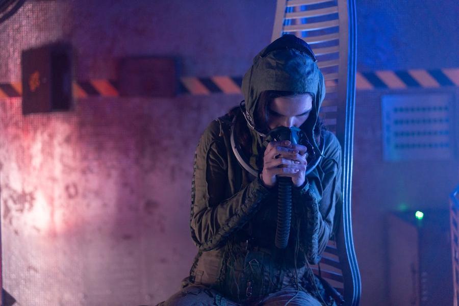 Man in a green military jacket, hunched over in front of a large, metal vehicle.
