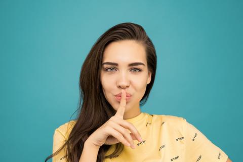 A female-presenting person in a yellow shirt holds a shushing finger in front of their mouth.
