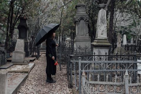 A woman dressed in black, holding a black umbrella and roses, standing in front of a tombstone.
